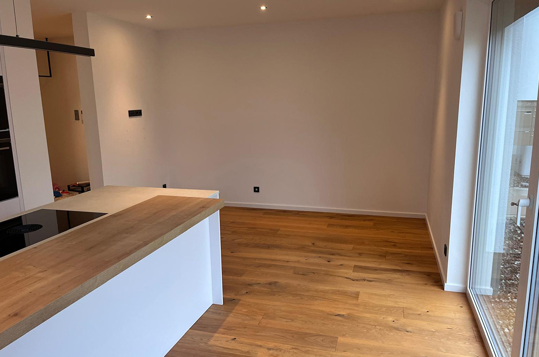 Leerer heller Wohnraum mit Holzboeden und weisser Kuecheninsel am Fenster