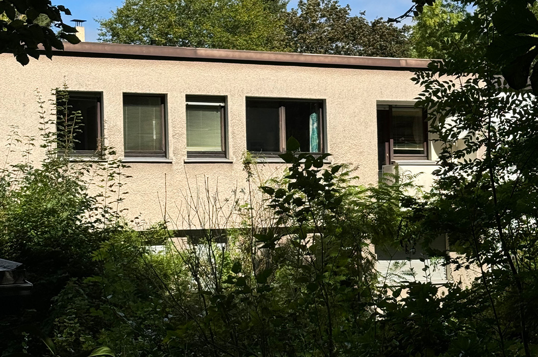 Beiges Haus mit Fenstern, dunklen Rahmen und gruenem Buschwerk im Vordergrund
