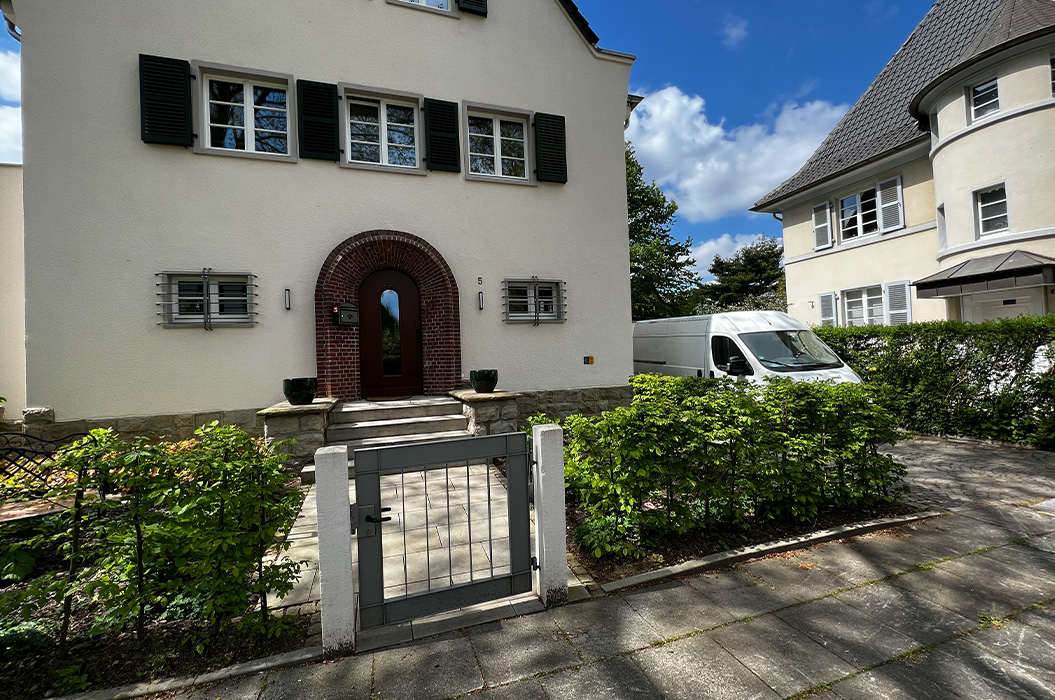 Beiges Haus mit Tor, ernstem Eingangsbogen, grüner Hecke und weißem Van