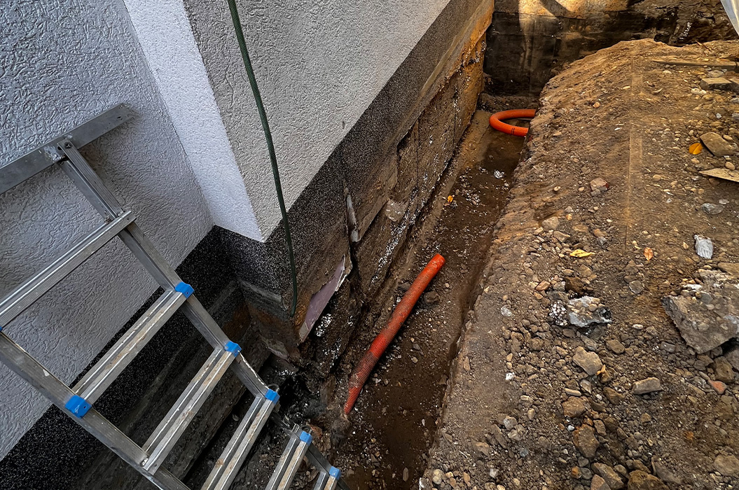 Bauarbeiterloser Graben neben Hauswand mit rotem Rohr und Leiter