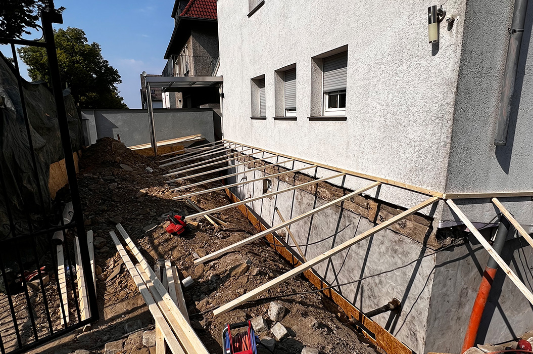 Baustelle am grauen Haus, Holzstreben im Graben, sonnig, rotbraunes Dach