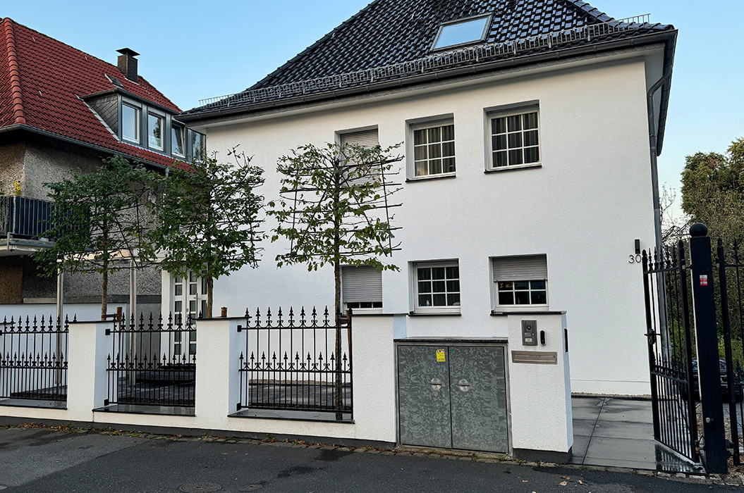 Weisses Wohnhaus mit dunklem Dach hinter Zaun und jungen Baeumen