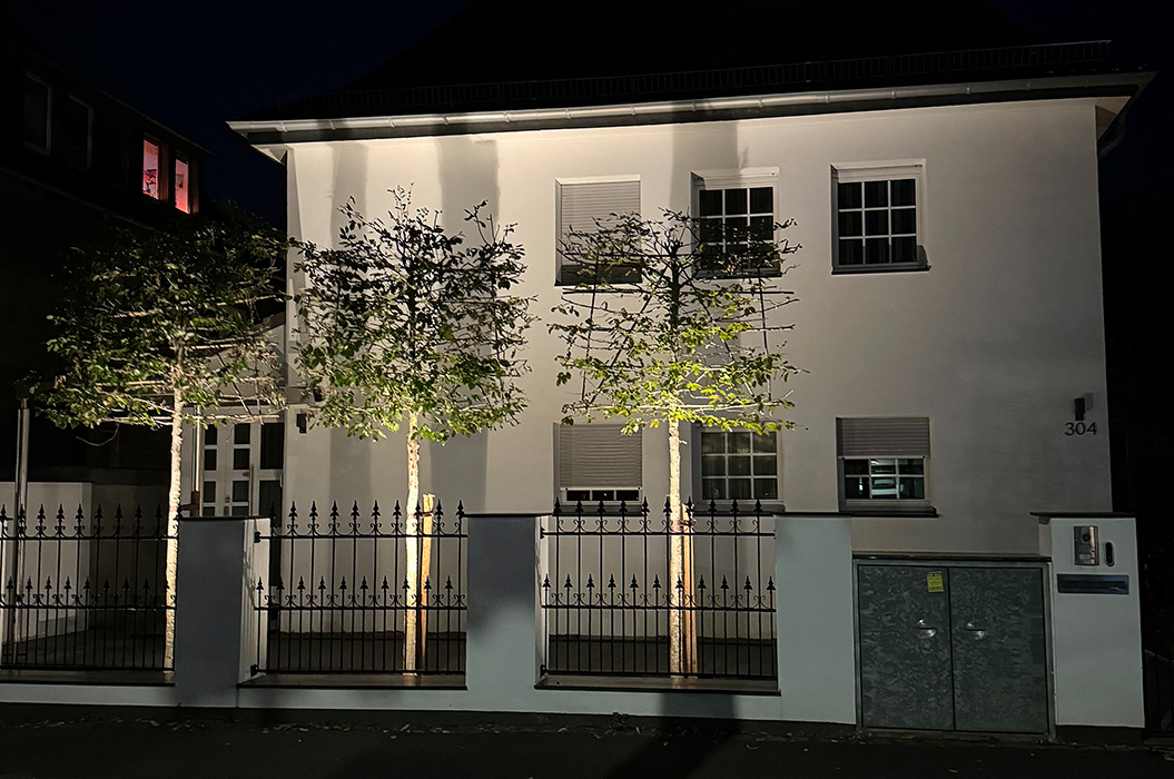 Beleuchtetes weisses Haus nachts mit Zaun, drei Baeumen und Fensterreihe